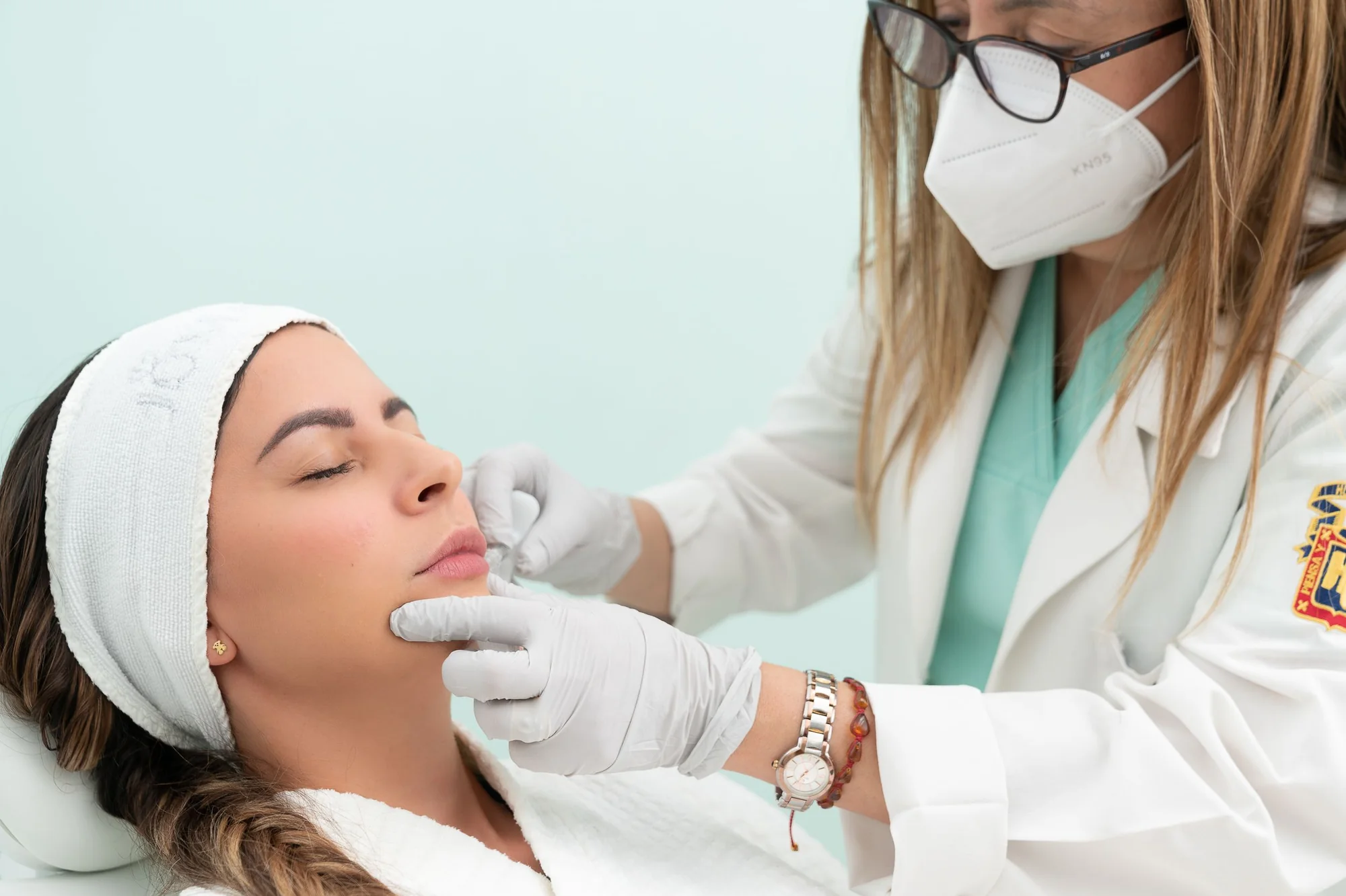 Especialista con mascarilla revisa el rostro de una paciente recostada antes de un tratamiento facial